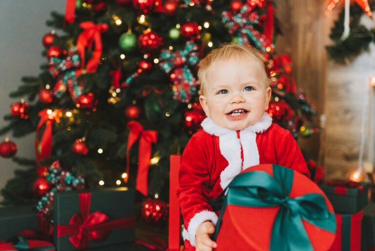 A smiling toddler dressed in a red Santa outfit holds a round gift box in front of a beautifully decorated Christmas tree with red and green ornaments and glowing lights.