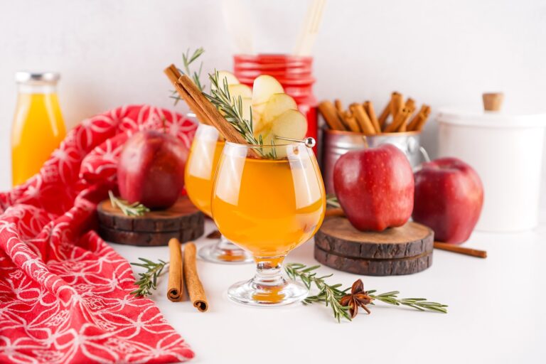 Festive Fireball apple cider cocktails garnished with apple slices, rosemary, and cinnamon sticks surrounded by apples and holiday spices.