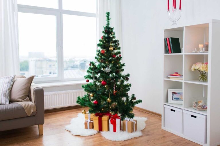 A decorated artificial Christmas tree stands in a bright living room, adorned with red and gold ornaments, candy canes, and warm string lights. Wrapped gifts with bows sit on a white fluffy tree skirt below. A sofa, large window with sheer curtains, and a white shelving unit with books, candles, and flowers complete the cozy holiday setting.