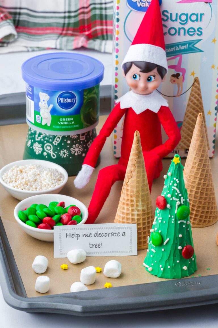 An Elf on the Shelf doll sits on a baking tray next to sugar cones and ingredients for decorating an edible Christmas tree. One cone is fully decorated with green frosting, red and green M&M candies, and white sprinkles to look like a Christmas tree, while other plain cones stand nearby. Small bowls of sprinkles and M&M candies, mini marshmallows, yellow star sprinkles, and a note reading “Help me decorate a tree!” are arranged around the elf. A tub of green vanilla frosting and a box of sugar cones are visible in the background with a festive plaid blanket behind them.