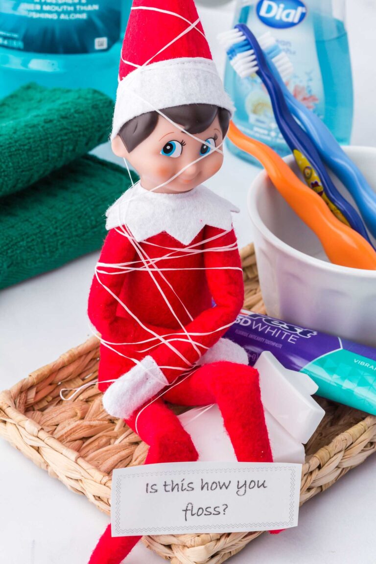 An Elf on the Shelf doll is tangled up in dental floss, wrapped around its head and body, sitting in a small bathroom basket. Nearby are toothbrushes, toothpaste, and mouthwash, along with a note that reads, “Is this how you floss?” creating a silly oral-care-themed prank.