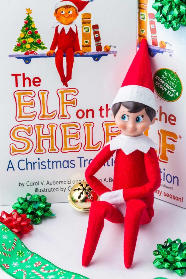 A classic Elf on the Shelf doll wearing a red felt outfit and pointed hat sits with crossed legs in front of the official The Elf on the Shelf: A Christmas Tradition book. The background shows the illustrated book cover featuring a small elf sitting on a shelf next to a Christmas tree and books. Festive green and red gift bows, a gold jingle bell, and a green candy-cane ribbon lie around the elf, creating a cheerful holiday scene.