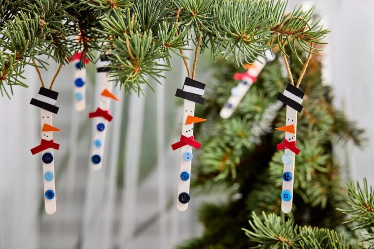Three handmade popsicle stick snowman ornaments hang from the branches of a green pine tree. Each snowman is crafted from a white-painted wooden stick decorated with small blue buttons, a red paper scarf, a tiny orange carrot-shaped nose, black marker eyes and coal dots, and a black paper top hat with a white band. The ornaments hang by simple twine loops, adding a charming homemade touch to the Christmas tree.