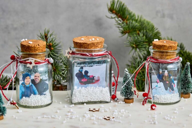 Three upcycled glass jars are turned into DIY photo snow globes, each filled with small white foam beads to mimic snow and featuring a family winter photograph inside. The jars are topped with cork lids and decorated with red and white twine, tiny wooden stars, and small festive beads. A pair of miniature bottle-brush Christmas trees sits in front of the jars, with a blurred evergreen branch in the background adding to the cozy holiday scene.
