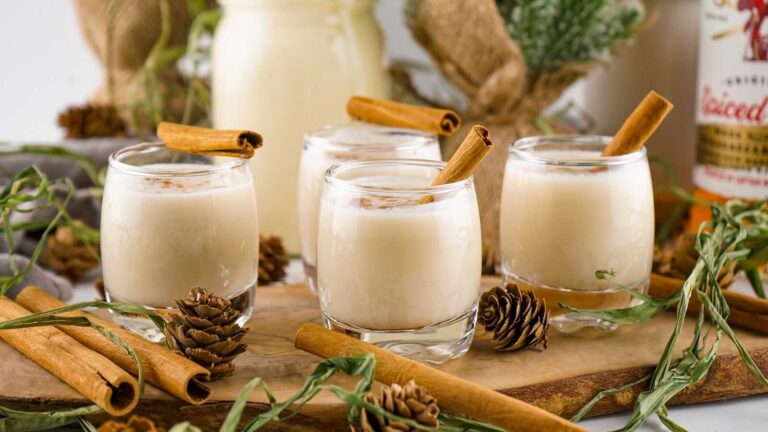 Eggnog shots with cinnamon sticks displayed on a wooden board with pinecones and holiday decor.
