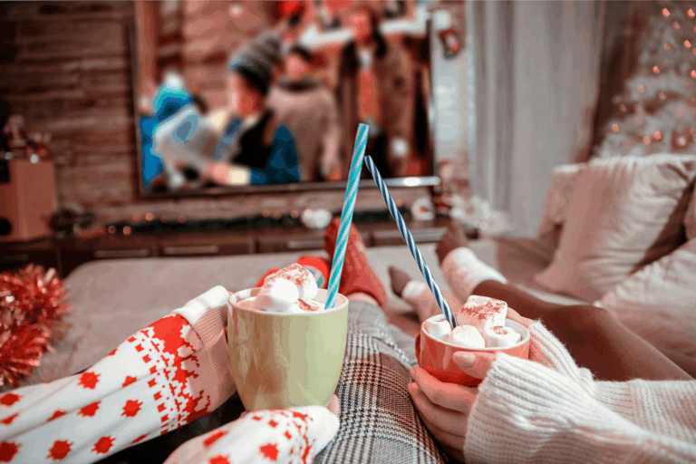 Cozy holiday movie night with two people holding mugs of hot chocolate topped with marshmallows.