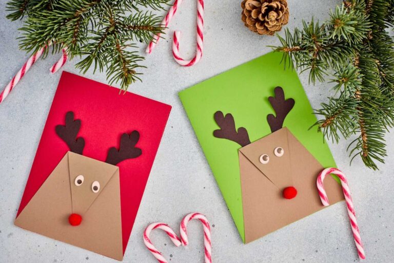 Two handmade reindeer Christmas cards sit side by side—one on a red background and one on a green background. Each card is made from folded brown cardstock forming a triangular reindeer face, decorated with dark brown paper antlers, googly-style paper eyes, and a small red pom-pom nose. Candy canes, pine branches, and a pinecone surround the cards, creating a festive holiday scene.