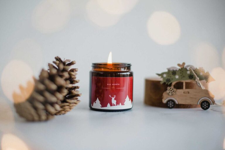 A lit holiday-themed soy candle in a brown glass jar with a red label featuring white silhouettes of trees and a deer sits on a white surface. To the left is a large pinecone, and to the right is a small wooden toy car carrying a green Christmas tree on its roof. Soft, warm bokeh lights blur in the background, creating a cozy holiday atmosphere.