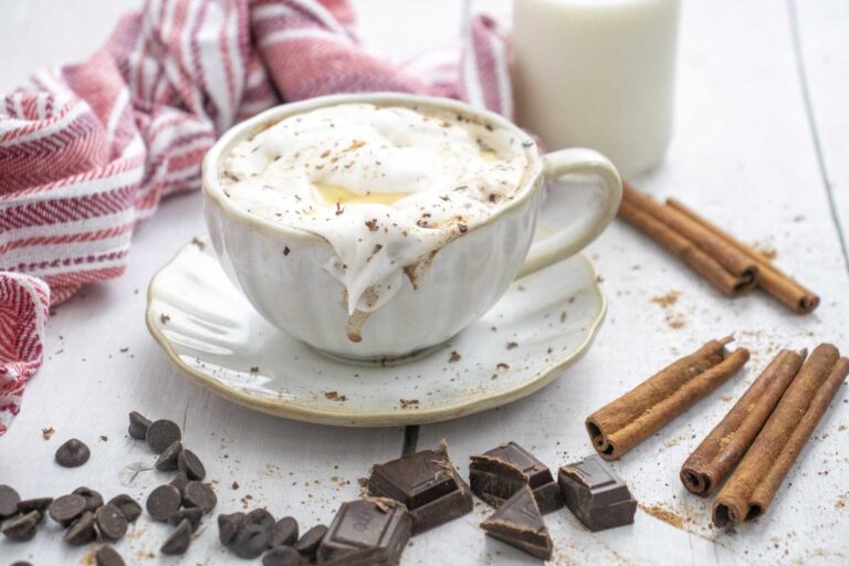 Cup of eggnog hot chocolate topped with whipped cream, surrounded by cinnamon sticks and chocolate.