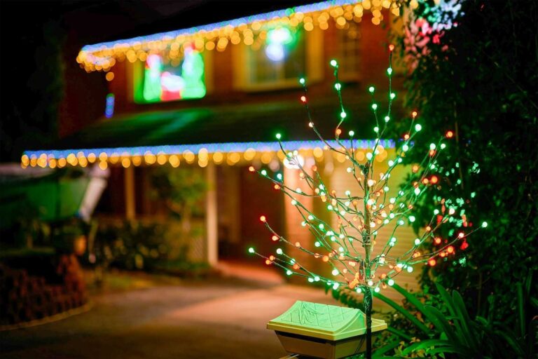 Light-up decorative tree with multicolor bulbs in front of a house with glowing Christmas lights.