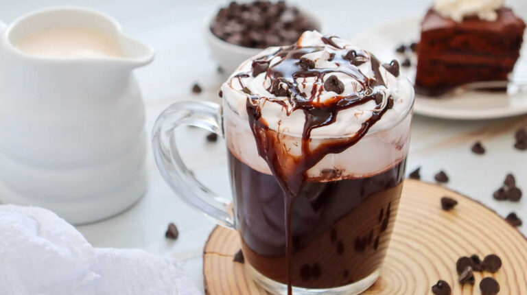 Starbucks copycat mocha hot chocolate with whipped cream, chocolate drizzle, and chocolate chips in a glass mug on a wooden board.