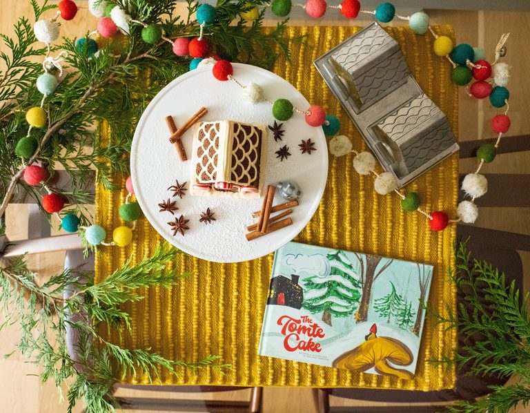 Flat lay of The Tomte Cake storybook, gingerbread house baking pan, Tomte gnome, and a decorated gingerbread cake with cinnamon sticks.